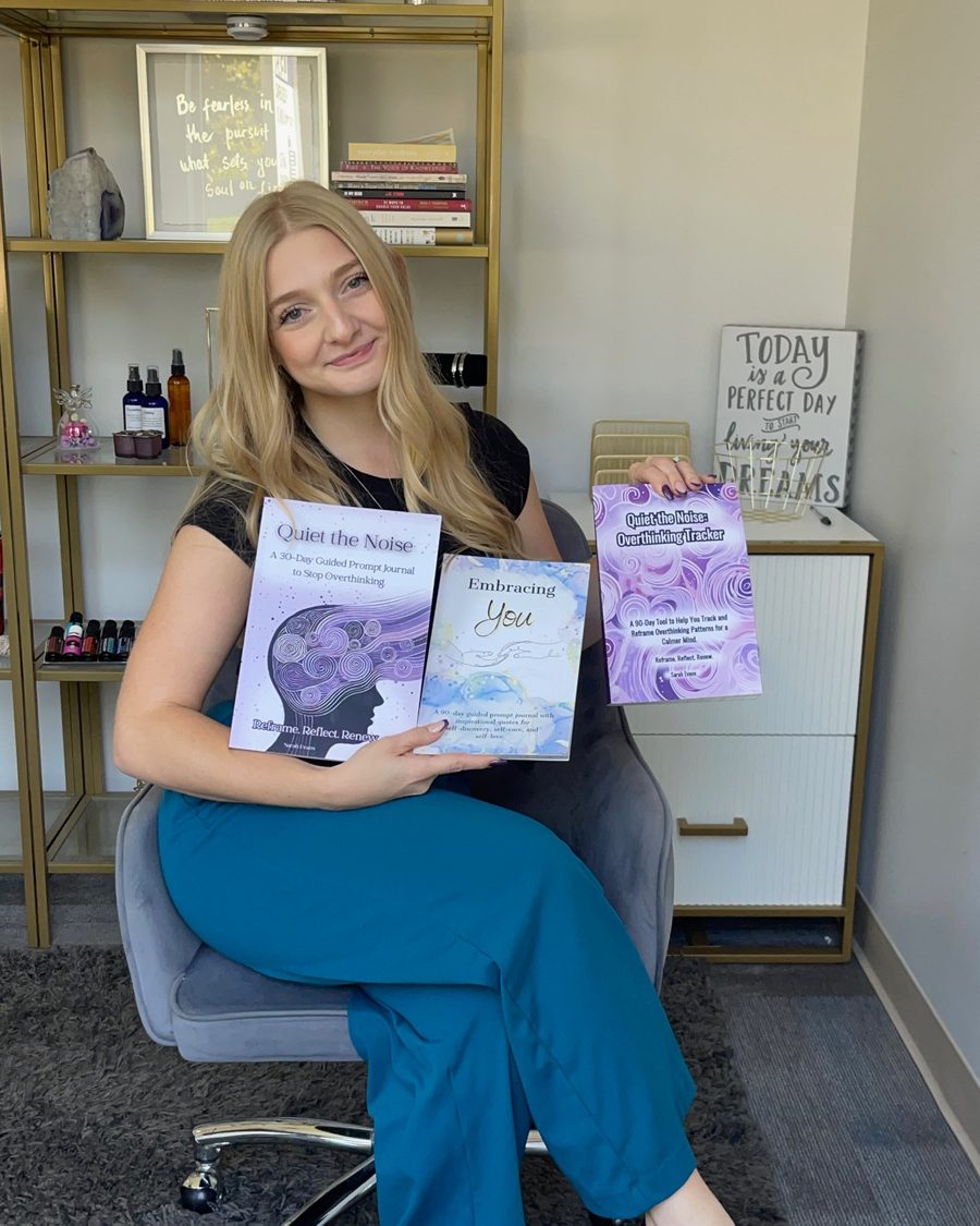 Sarah Evans in her office, holding her journals Quiet the Noise, Embracing You and the Overthinking Tracker